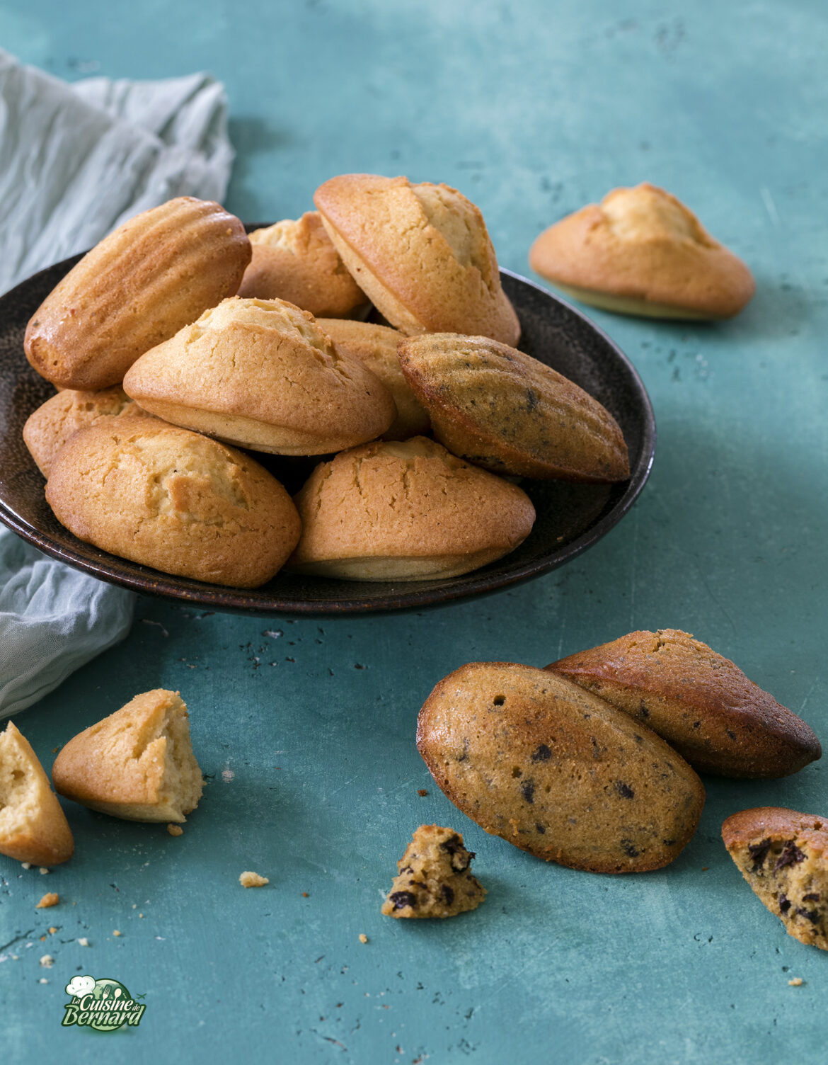 Une assiette de madeleines dorées, confectionnées pour réaliser ce qui semblait impossible, repose sur une surface bleue. Quelques morceaux cassés sont éparpillés un peu partout, avec une serviette bleue à proximité.