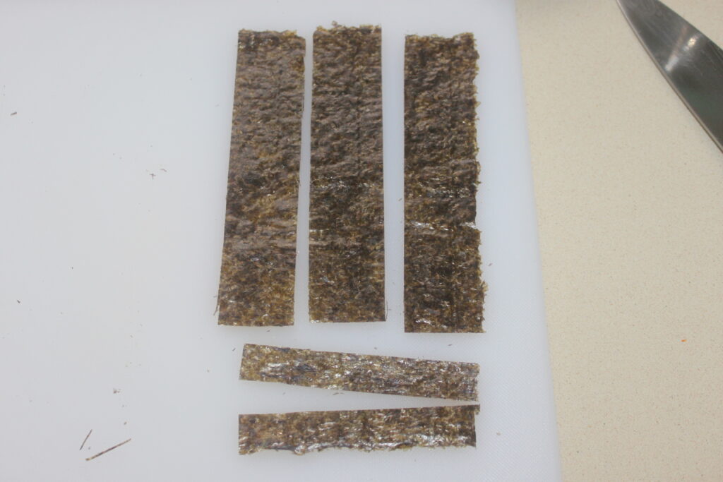 Quatre longues bandes rectangulaires et deux courtes d'algues sur une planche à découper blanche. Un couteau est partiellement visible à droite.