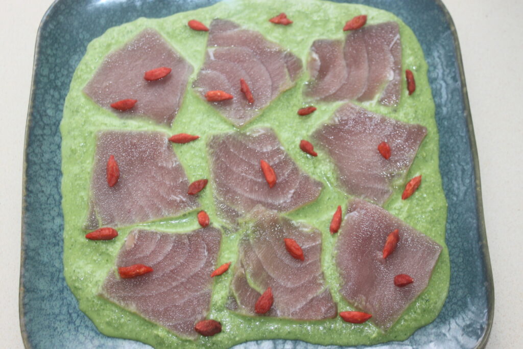Sept tranches de poisson cru disposées sur une purée verte, garnies de petites baies rouges, servies sur une assiette carrée bleu vert évoquent l'essence du ceviche de thon.