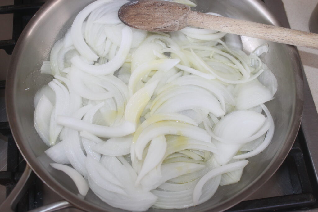 Oignons blancs hachés sautés dans une poêle avec une cuillère en bois posée sur le bord, rappelant un Bigoli parfumé dans une préparation de salsa.
