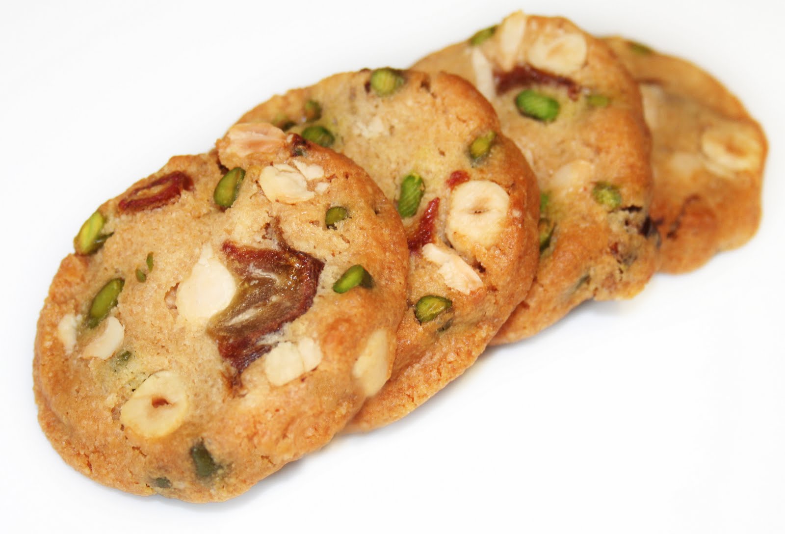 Quatre sablés ronds avec des morceaux visibles de noix et de fruits secs sont placés sur une seule rangée sur un fond blanc.