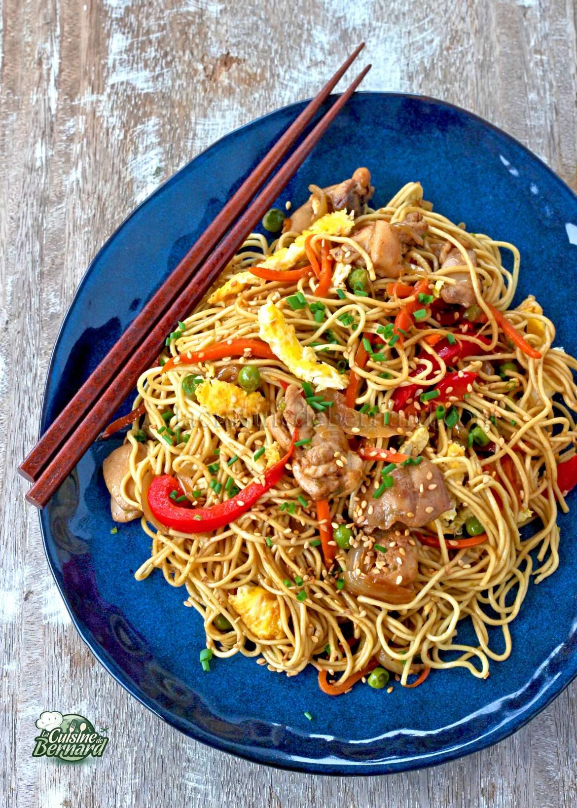 Une assiette bleue contient une portion de nouilles sautées avec des légumes et des morceaux de viande, garnies de graines de sésame et accompagnées d'une paire de baguettes.