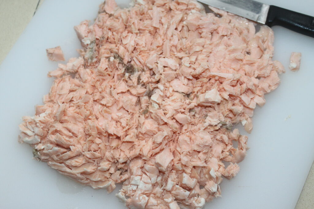 Saumon cuit haché sur une planche à découper blanche avec un couteau à proximité, prêt à compléter les raviolis crémeux à la ricotta.