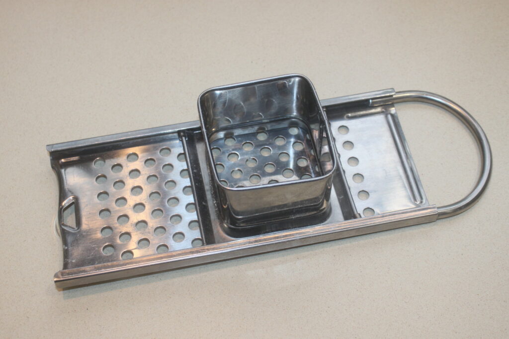 Râpe en métal avec manche et récipient carré perforé, parfaite pour râper des ingrédients comme les Spätzles, sur une surface beige.