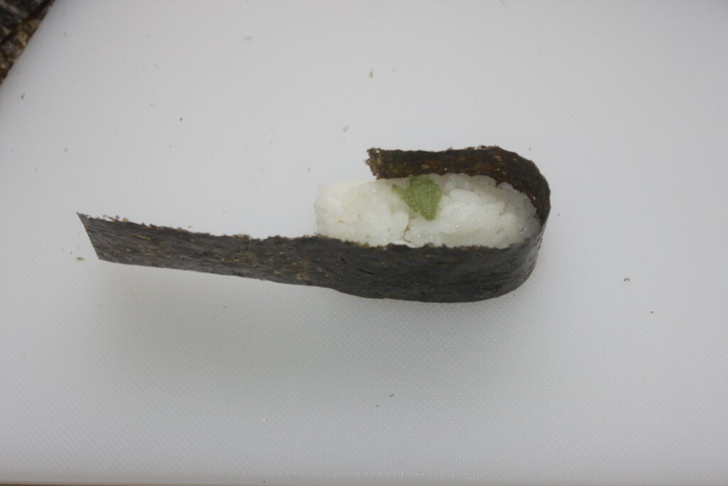 Un morceau de sushi avec du riz, du wasabi et des algues, partiellement roulé sur une surface blanche.