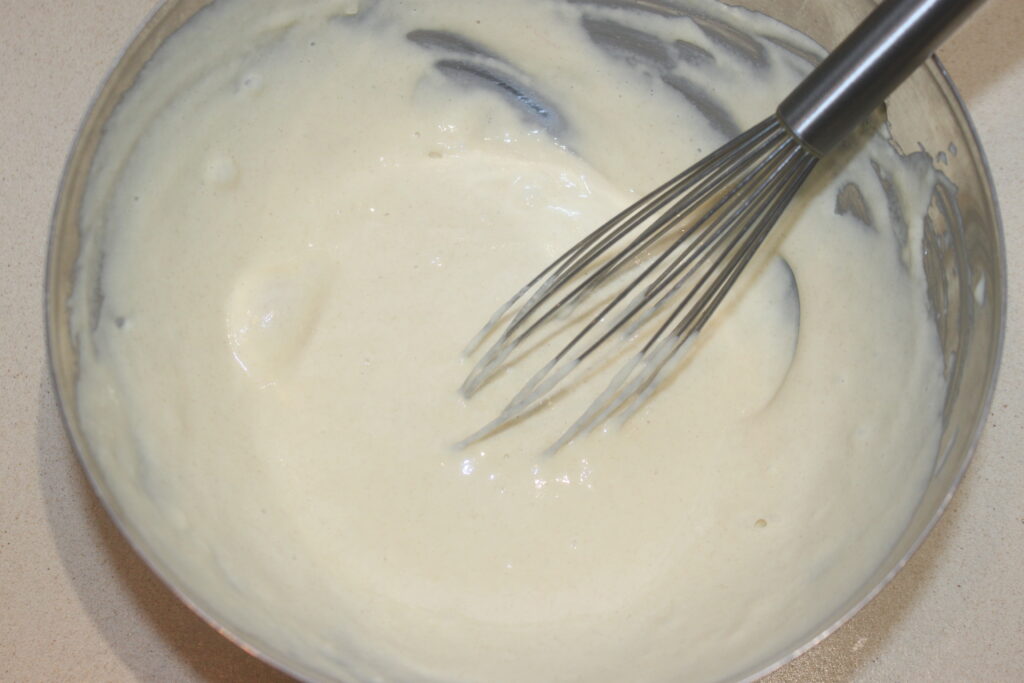 Un fouet en métal est posé sur le plan de travail dans un bol de pâte lisse et claire, prêt à créer de délicats Spätzles.