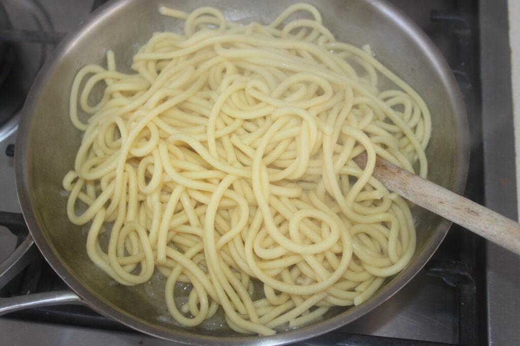 Les pâtes bucatini et la salsa salée mijotent ensemble dans une casserole en acier inoxydable, remuées doucement avec une cuillère en bois sur la cuisinière.