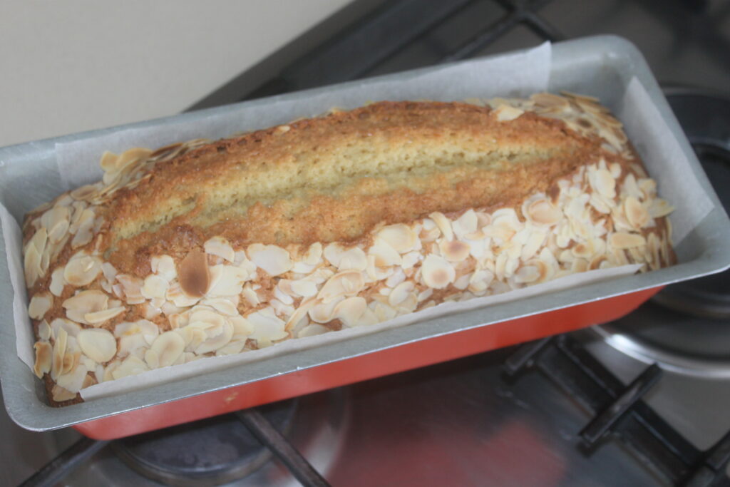 Une miche de gâteau mégamande rectangulaire avec des amandes tranchées sur le dessus, cuite dans un moule en métal placé sur une cuisinière.
