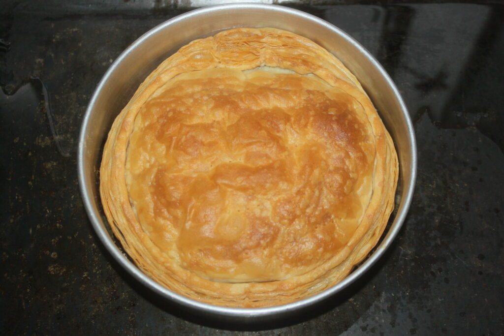 Une pâte feuilletée dorée, rappelant une tarte tatin classique, se présente joliment dans un moule à pâtisserie rond en métal sur une surface sombre.