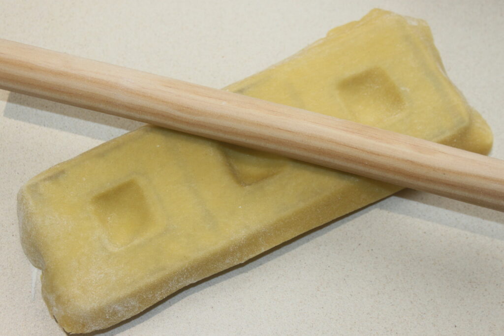 Un rouleau à pâtisserie en bois repose sur une feuille de pâte jaune sur une surface claire, prêt à confectionner de délicieux raviolis.