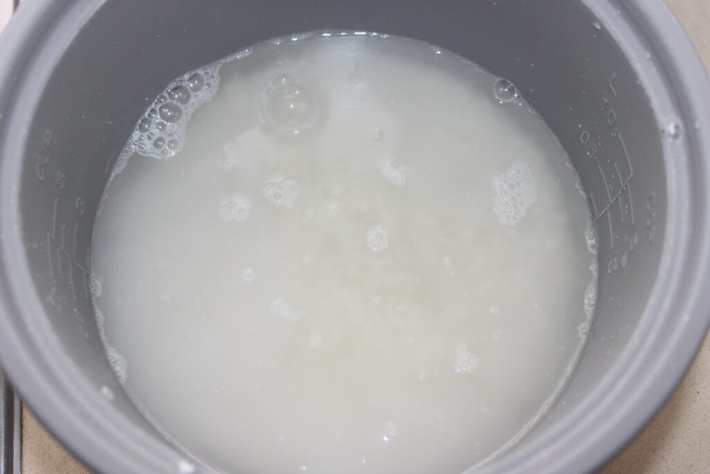 Gros plan du riz blanc en cours de cuisson dans un cuiseur à riz gris, avec de l'eau et des bulles visibles.