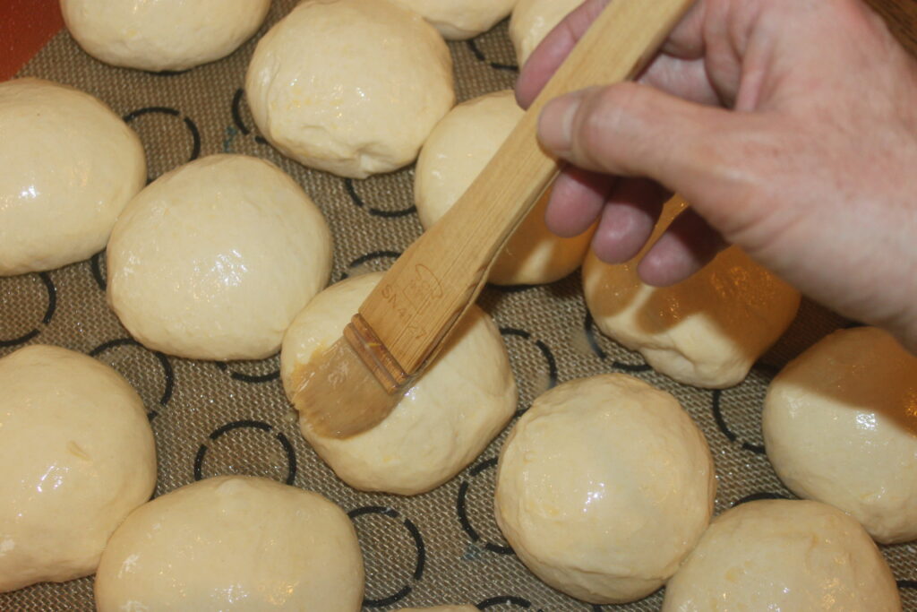 Une main badigeonne délicatement d'œuf battu les petits pains moelleux, des boules de pâte parfaitement rondes posées sur un tapis en silicone.