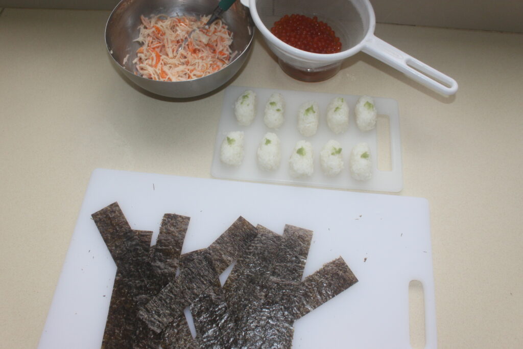 Ingrédients pour la préparation des sushis : feuilles d'algues nori, boulettes de riz au wasabi, crabe râpé et caviar rouge, disposés sur une surface blanche.