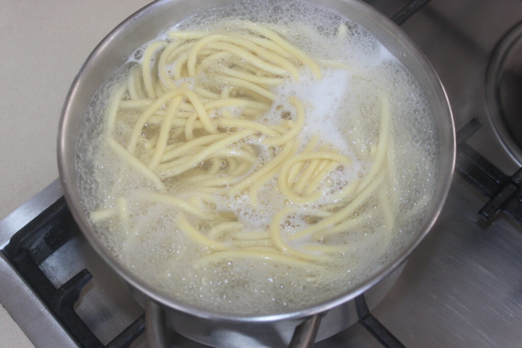 Un plat de pâtes traditionnel, les Bigoli en salsa, est en train de bouillir dans une casserole en métal sur la cuisinière, avec de l'eau bouillonnante et de la mousse visible à la surface.