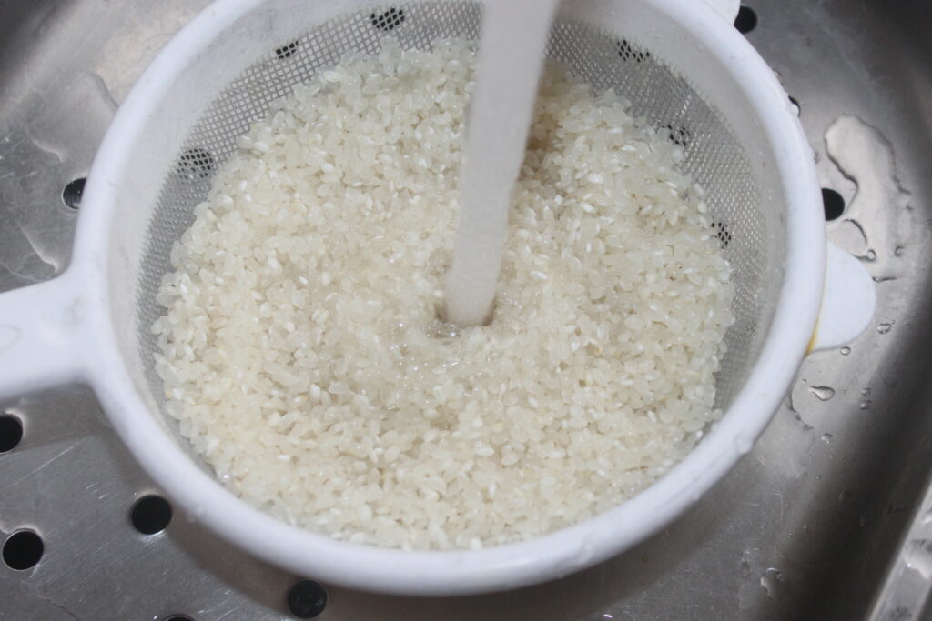 Riz rincé sous l'eau courante dans une passoire blanche placée au dessus d'un évier.