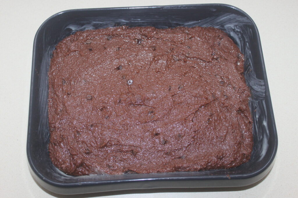 Un moule carré rempli de pâte à brownie au chocolat non cuite saupoudrée d'amandes croquantes repose sur un plan de travail clair.