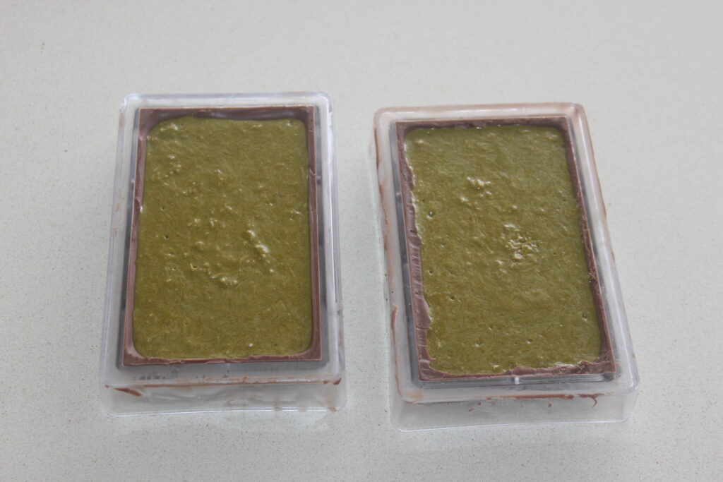 Deux moules rectangulaires remplis d'un mélange vert, partiellement entourés d'une couche de chocolat de luxe, sont placés sur un plan de travail de couleur claire.