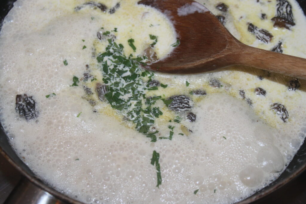 Gros plan d'une sauce à la crème bouillonnante dans une casserole, avec des morilles, du persil et du fromage râpé, le tout remué avec une cuillère en bois.