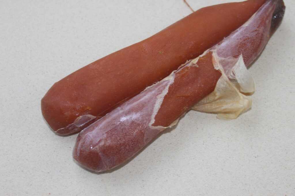 Deux morceaux d'œufs de poisson séchés reposent sur une surface légère, l'un partiellement enveloppé dans une enveloppe transparente, rappelant les saveurs délicates des raviolis saumon avec une touche de sauce aneth et citron vert.