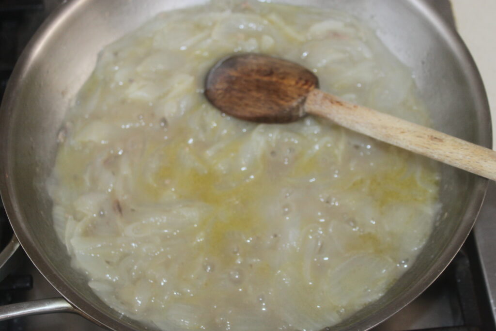 Une cuillère en bois repose dans une poêle d'oignons sautés nageant dans un liquide de couleur claire, rappelant un Bigoli dans une préparation de salsa, bouillonnant doucement sur la cuisinière.