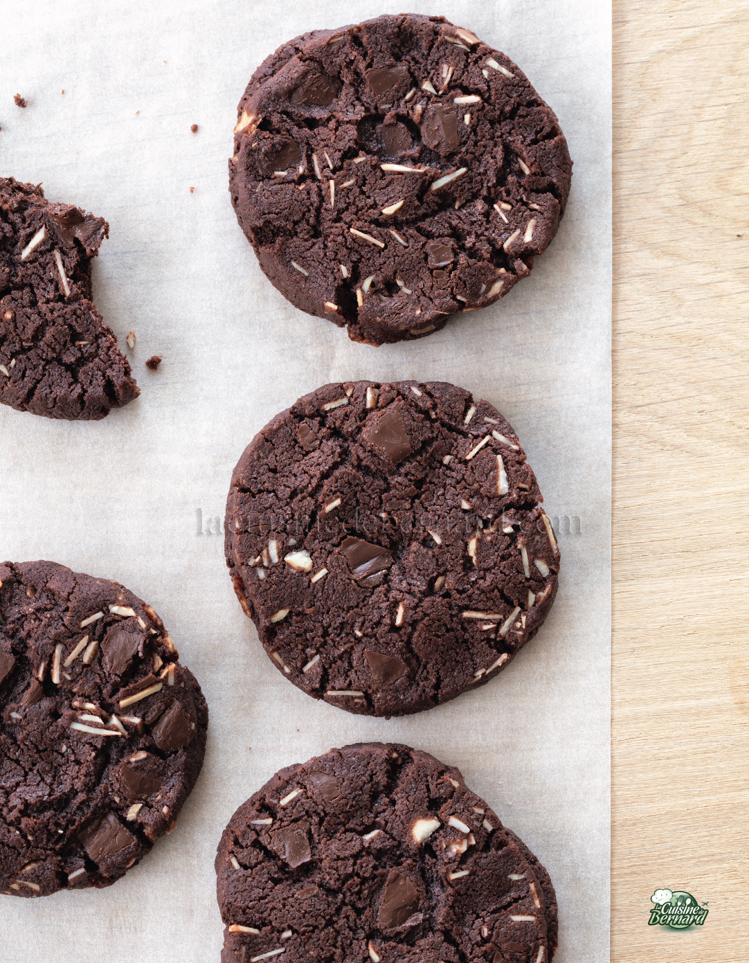 Quatre biscuits au chocolat entiers avec des morceaux de chocolat et des morceaux de noix sont disposés sur du papier sulfurisé Brouillon auto, avec une partie d'un biscuit cassé visible sur la gauche.