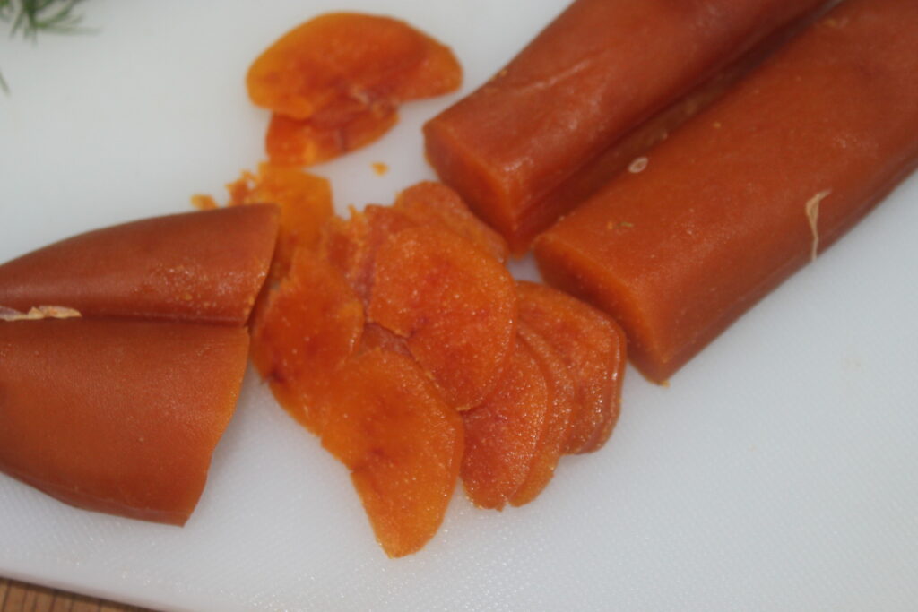 Des tranches d'œufs de poisson séchés reposent sur une planche à découper blanche, à côté de deux sections non tranchées, complétées par une pointe de citron vert pour un zeste supplémentaire.