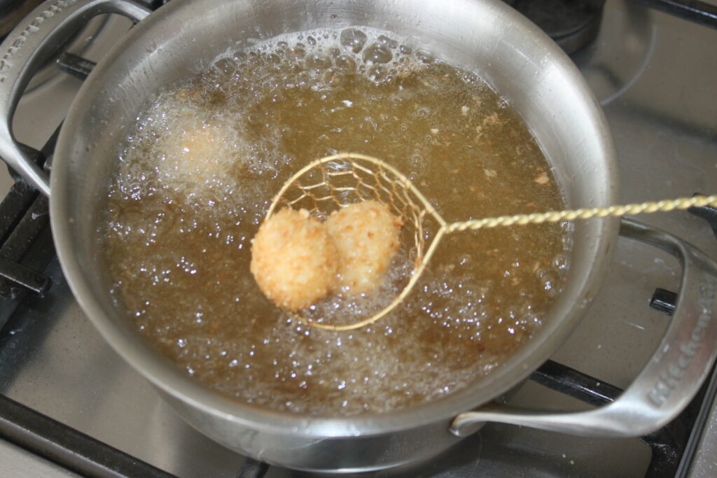 Una espumadera metálica levanta dos bolas de mozza empanada del aceite caliente de una sartén sobre una hornilla.