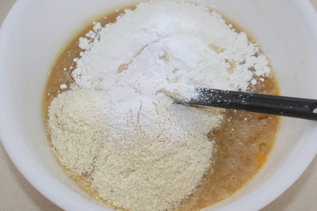 Un bol à mélanger contenant une pâte à gâteau brun clair est garni de tas de farine d'amande et de farine tout usage, avec une spatule noire partiellement immergée dans les ingrédients, créant une base parfaite pour votre dessert mégamande.