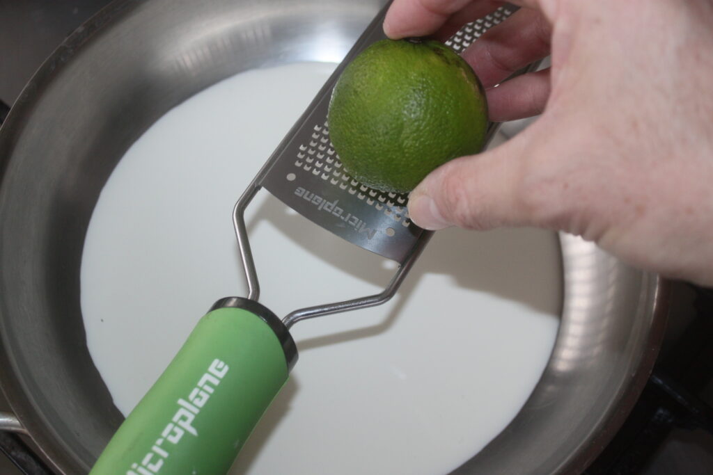 Râper à la main un citron vert au dessus d'une casserole remplie de lait à l'aide d'un zesteur à manche vert.