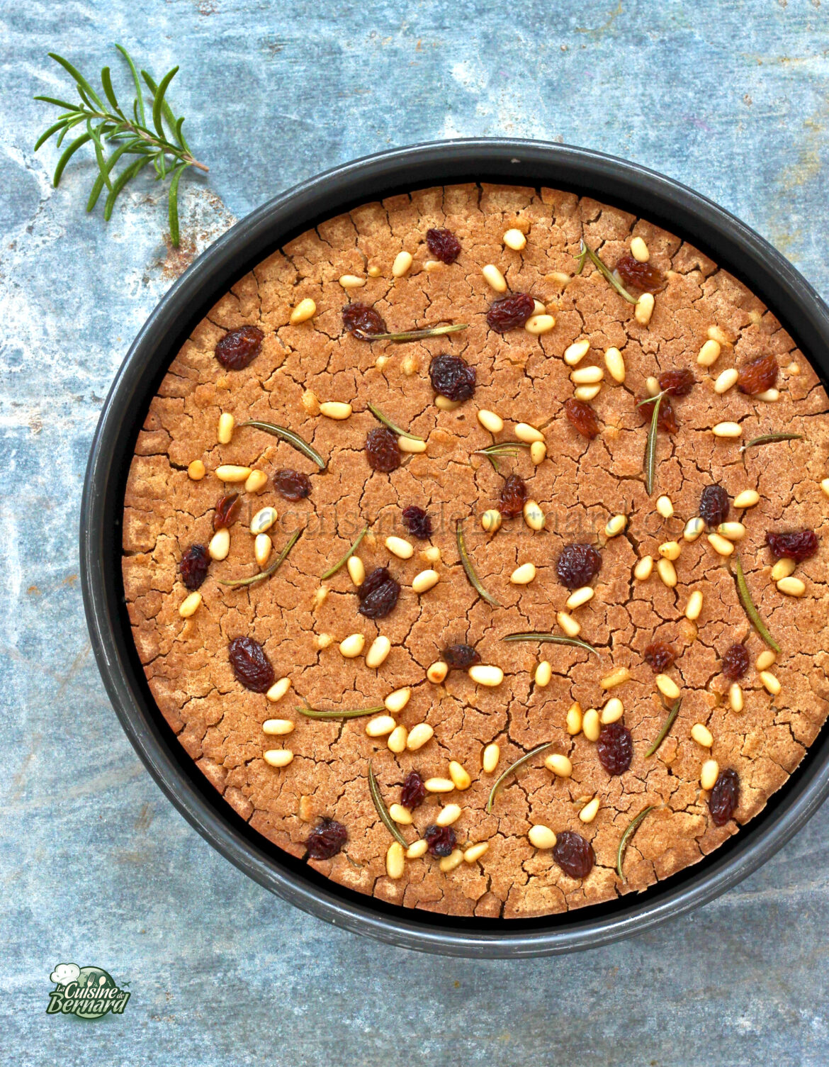 Un postre redondo, recién horneado, cubierto con piñones y romero, con pasas esparcidas en la superficie, presentado en un molde para hornear negro sobre un fondo con textura azul.
