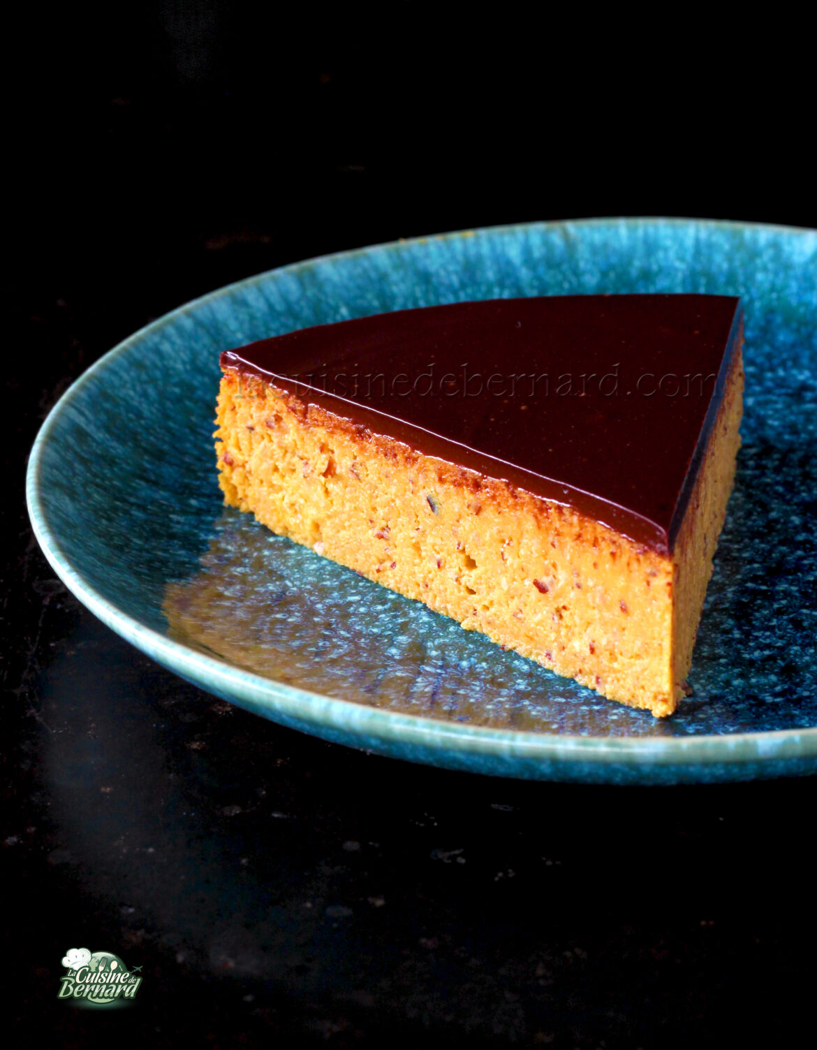 Une tranche de gâteau fondant à l'orange garnie de chocolat repose sur une assiette en céramique bleue sur fond sombre.