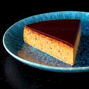 Une tranche de gâteau en couches avec une ganache au chocolat brillante sur une assiette bleue.