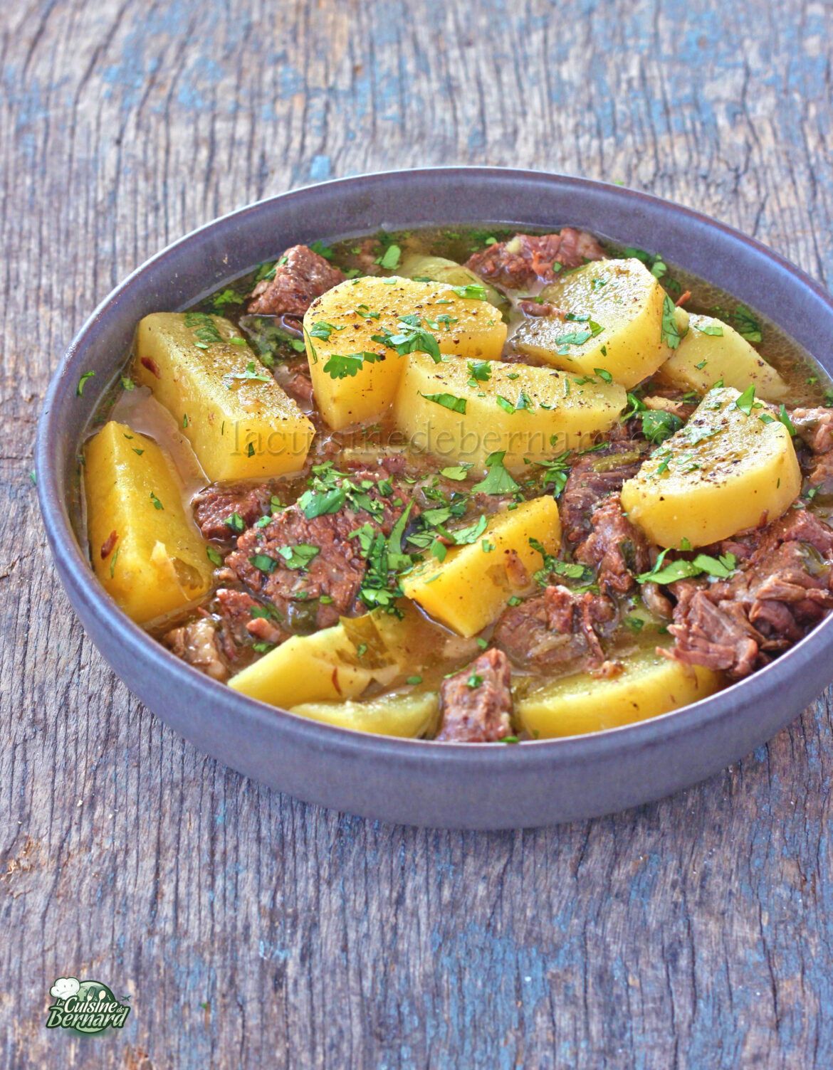 Un bol de ragoût de bœuf avec pommes de terre et garni de persil haché, servi sur une surface en bois.