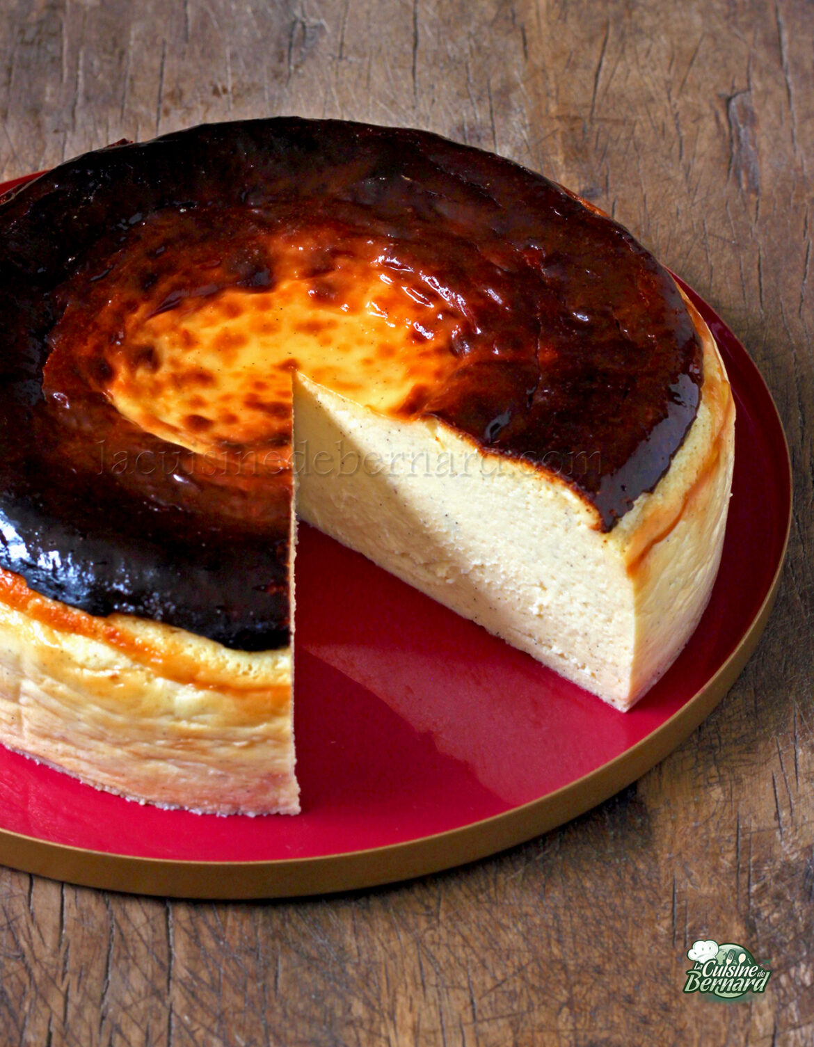 Un cheesecake basque brûlé entier dont une tranche a été retirée, révélant son intérieur crémeux, repose sur une assiette rouge sur une surface en bois.
