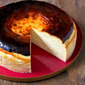 Una tarta de queso vasca redonda y quemada con la parte superior dorada se asienta sobre un plato rojo sobre una superficie de madera. Se corta una rebanada, dejando al descubierto un interior cremoso.