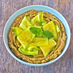 Un tazón de salsa cremosa adornado con rodajas de pepino y hojas de menta, colocado sobre una superficie con textura de madera.