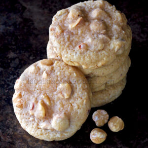 Una pila de galletas de nueces de macadamia con chocolate blanco junto a tres nueces de macadamia sobre una superficie oscura.