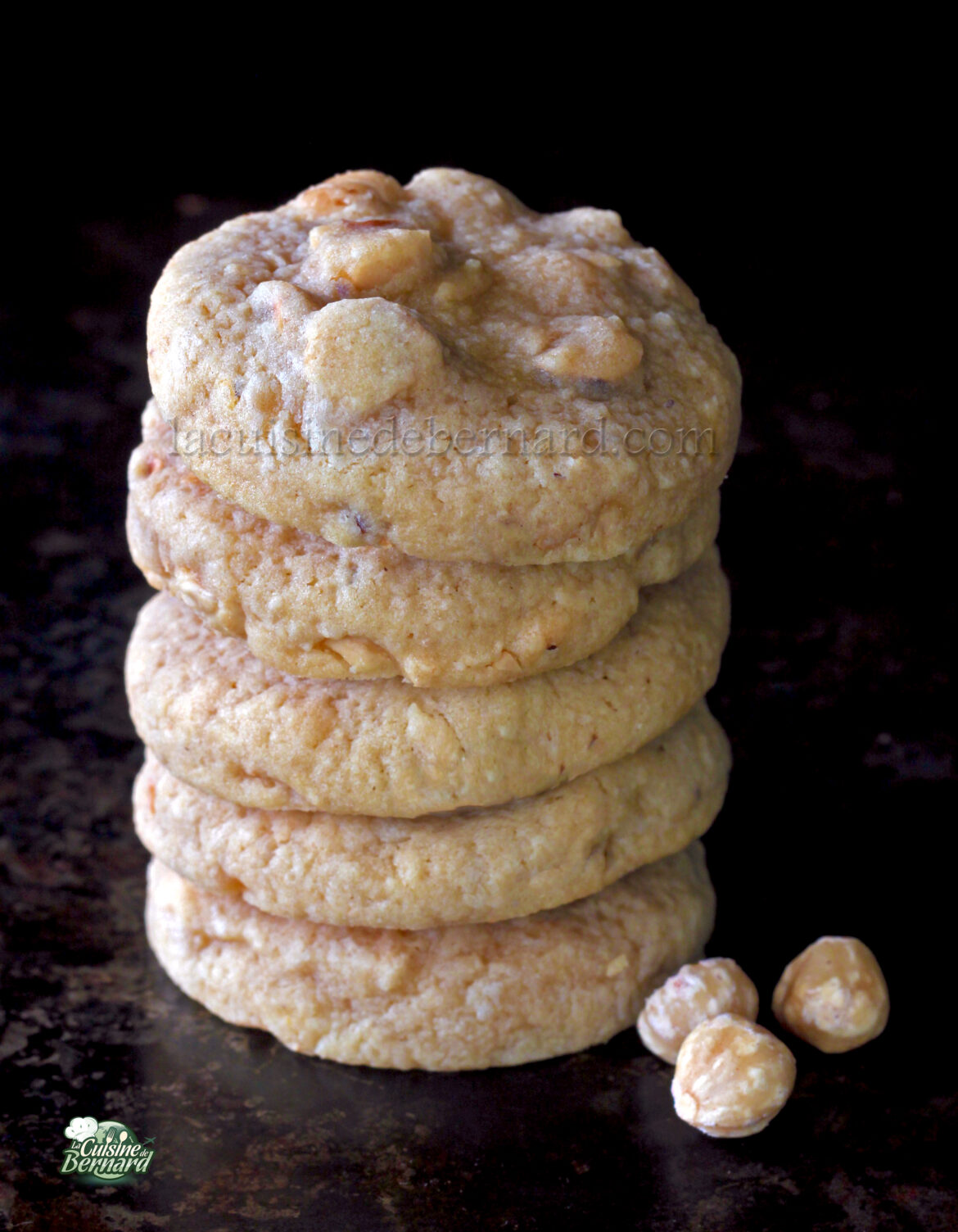 Une pile de cinq biscuits aux noisettes avec quelques noisettes éparpillées à la base, sur un fond sombre.