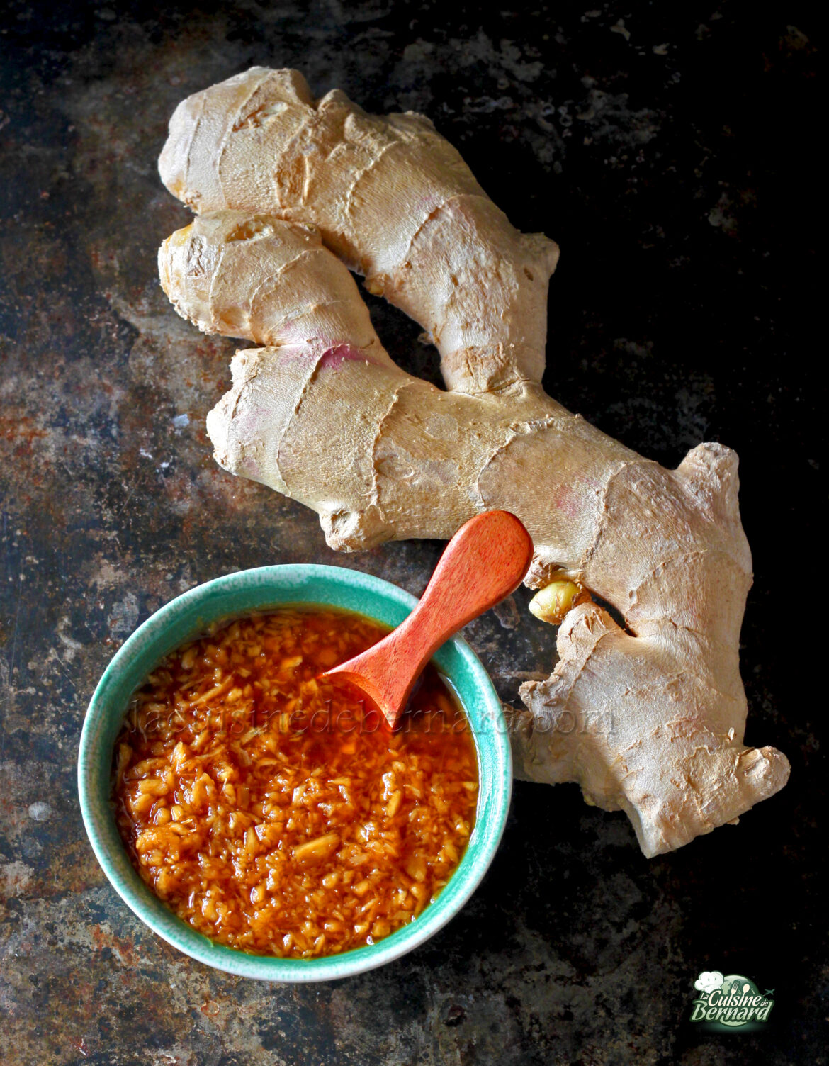 Un morceau de racine de gingembre frais se trouve à côté d'un bol de gingembre râpé dans une sauce brun rougeâtre, avec une cuillère en bois à l'intérieur du bol, le tout disposé sur une surface sombre.