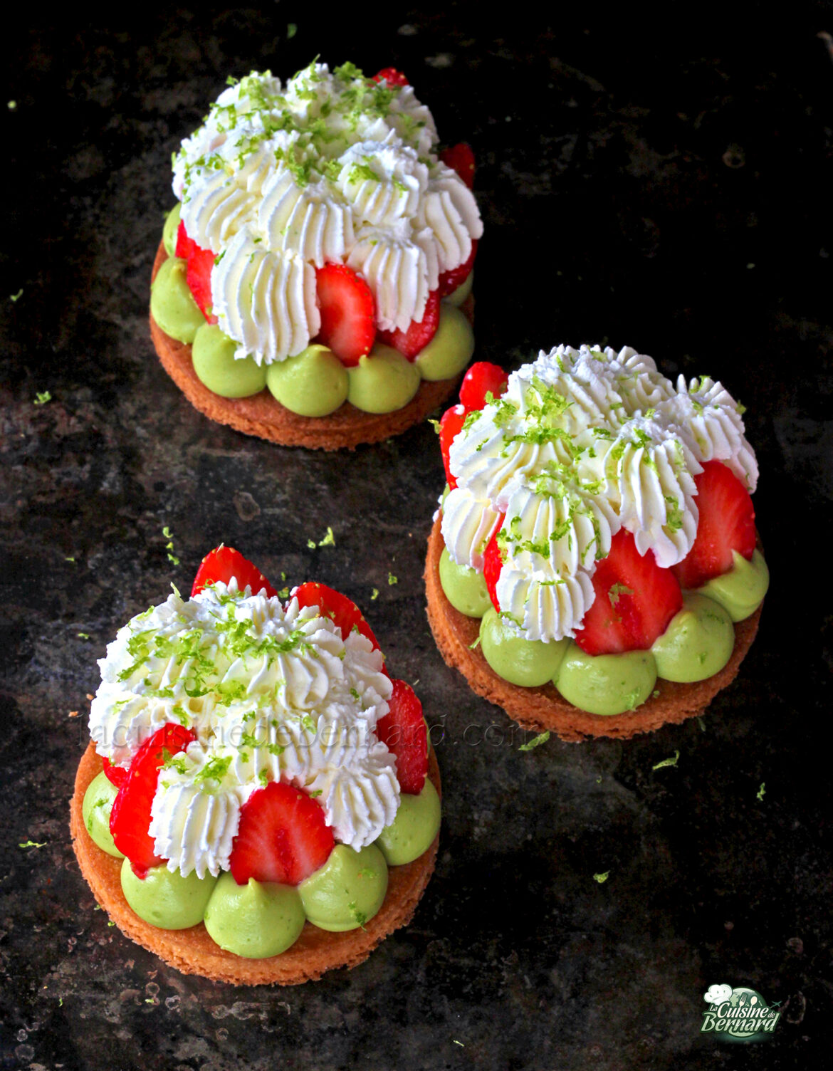 Trois Tartelettes fraîches individuelles garnies de mascarpone chantilly, de fraises, de crème verte et de zeste de citron vert sur une surface sombre.