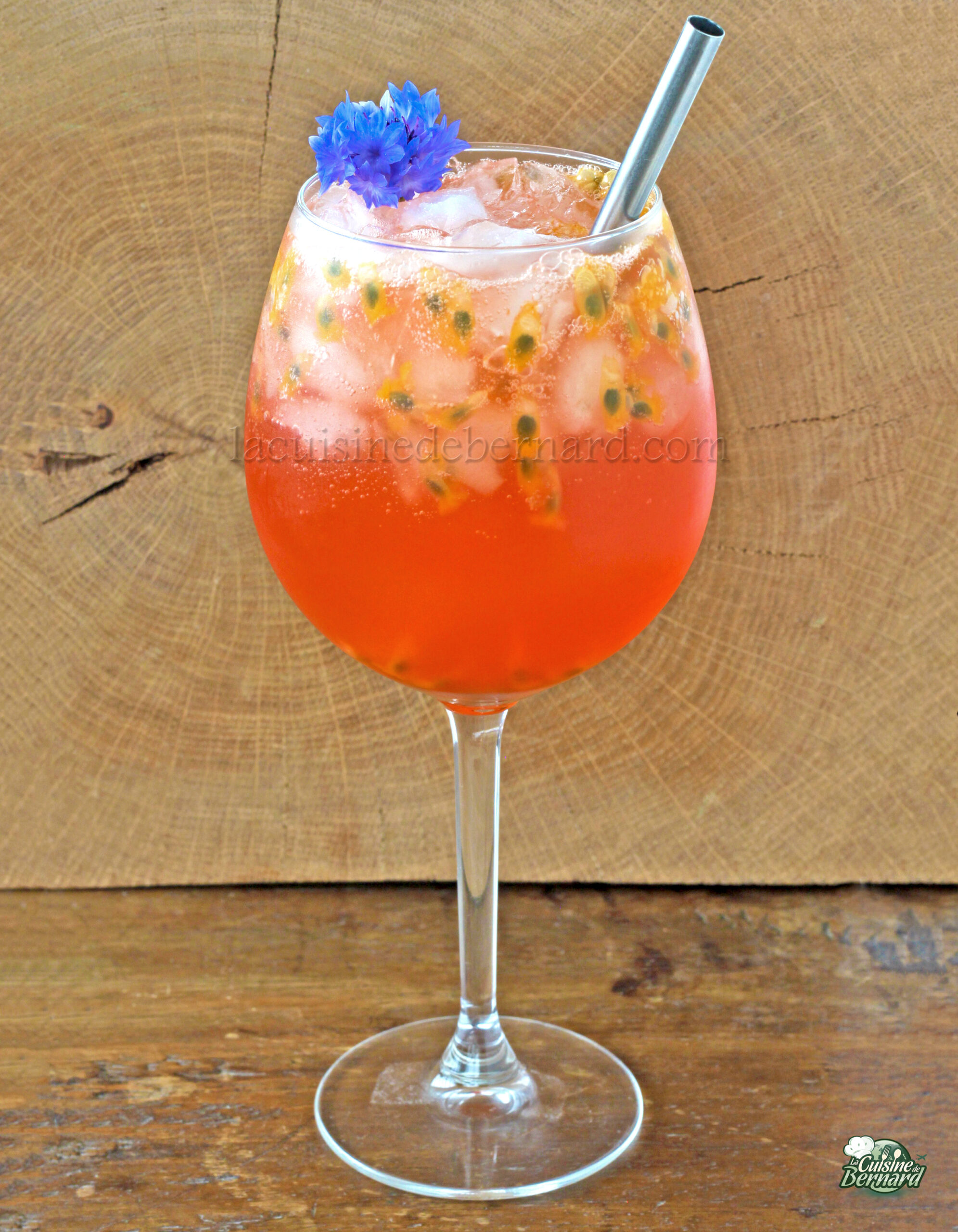 Un grand verre à pied rempli d'un cocktail fruité rouge vif garni d'une fleur violette et d'une paille en métal, sur un fond en bois, évoque le spritz parfait en bord de plage.