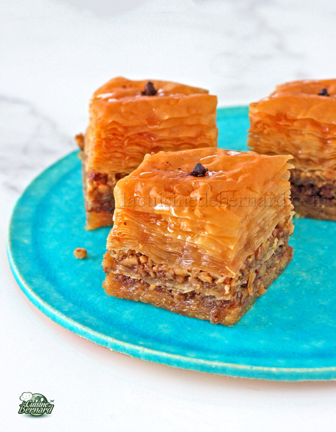 Gros plan de trois morceaux de baklava en couches avec noix sur une assiette turquoise. Chaque pièce est surmontée d'un clou de girofle, mettant en valeur les riches traditions des spécialités grecques.