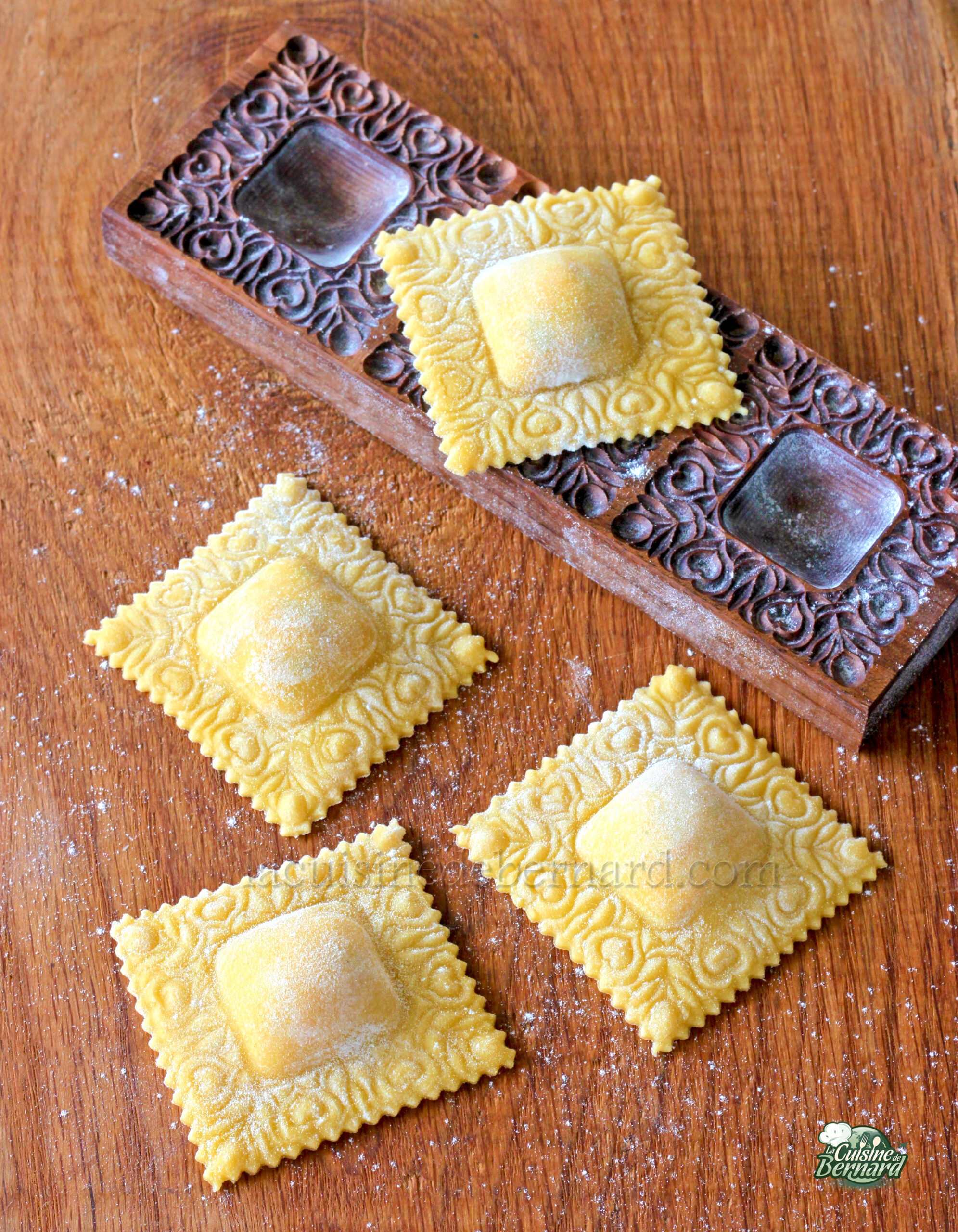 Quatre morceaux de raviolis carrés aux motifs décoratifs sur une surface en bois, à côté d'un moule en bois sculpté, sont soigneusement remplis de ricotta, ajoutant une texture crémeuse qui complète leur design délicat.