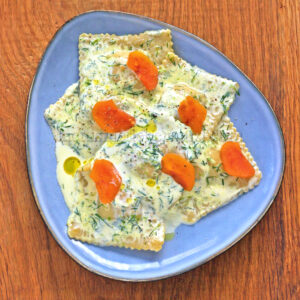 Une assiette de raviolis saumon nappés d'une sauce crémeuse aneth, agrémentés de tranches de carottes et d'un soupçon de citron vert, servis sur une table en bois.