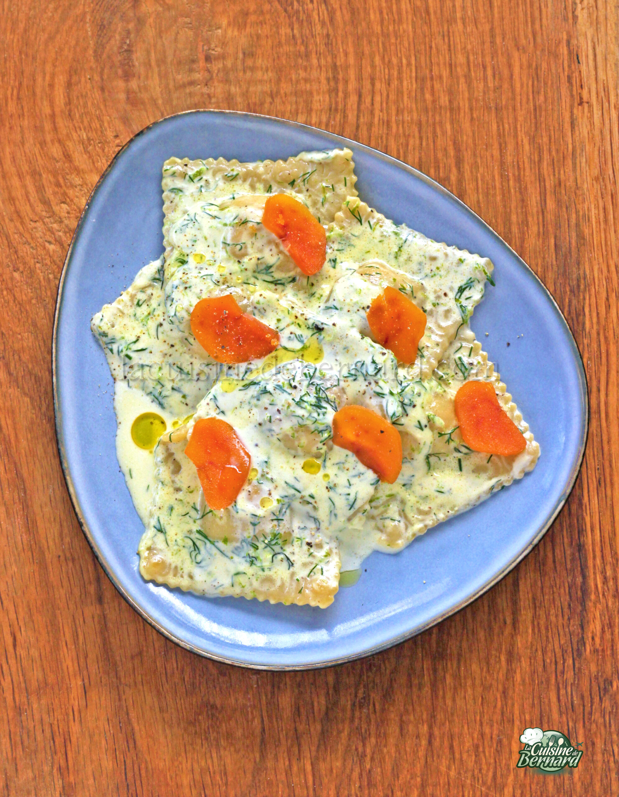 Une assiette de raviolis saumon nappés d'une sauce crémeuse aneth, agrémentés de tranches de carottes et d'un soupçon de citron vert, servis sur une table en bois.