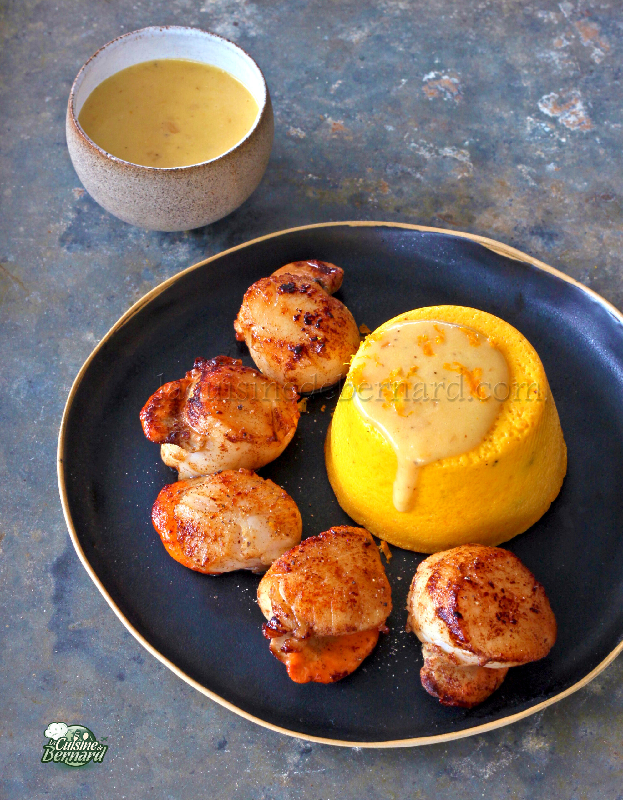 Des coquilles Saint Jacques grillées élégamment présentées sur une assiette noire, accompagnées de beurre à l'orange et d'un flan de potimarron vibrant. Une tasse de sauce supplémentaire trône en arrière plan, rehaussant l'attrait du plat.