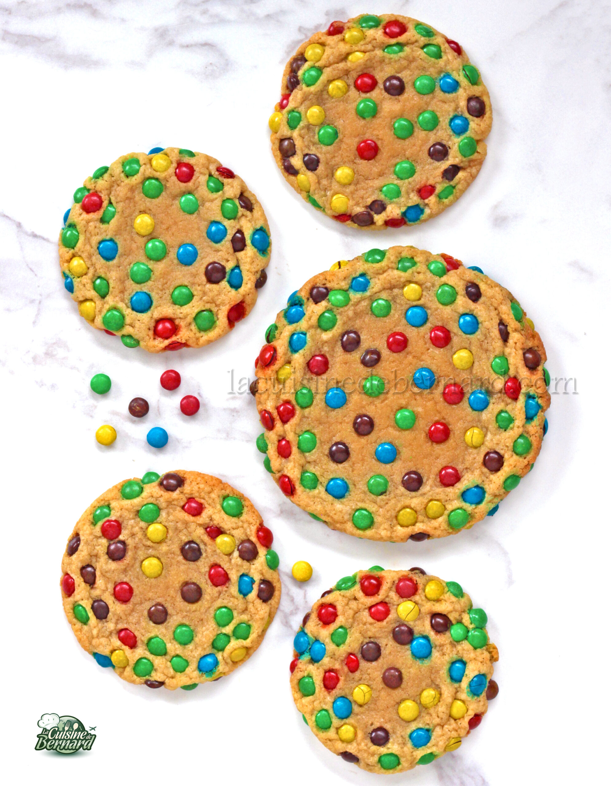 Six biscuits parsemés de mini M&M's vibrants sont dispersés sur une surface en marbre blanc, créant une délicieuse touche de couleur.