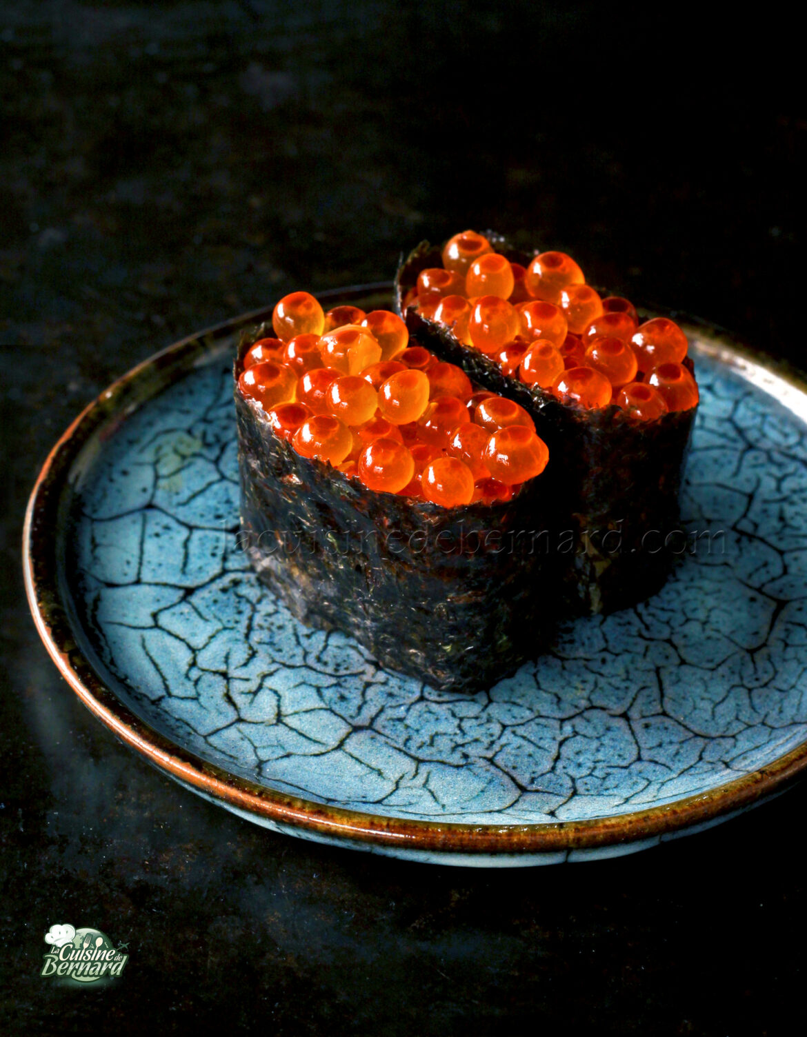 Deux morceaux de sushi garnis d'œufs de poisson orange vif sont servis sur une assiette bleue texturée.