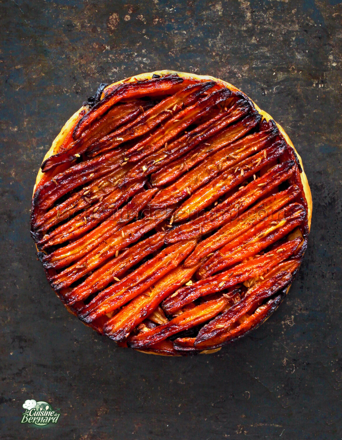 Une belle tarte tatin avec des couches caramélisées de carottes finement tranchées disposées soigneusement sur le dessus, présentées sur une surface texturée sombre.