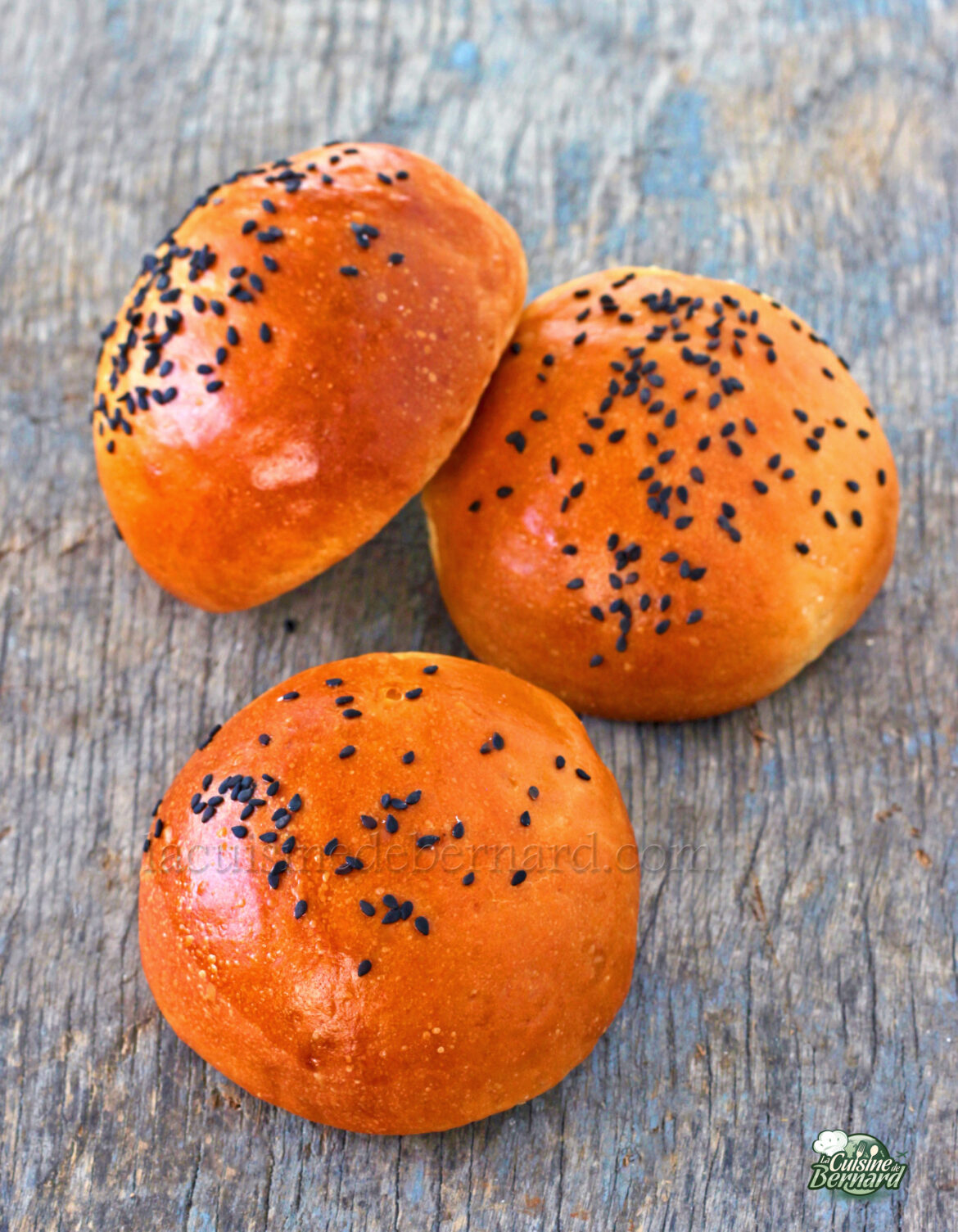 Trois petits pains moelleux dorés, garnis de graines de sésame noires, reposent élégamment sur une surface en bois.